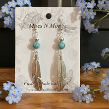 Load image into Gallery viewer, Feather-shaped earrings with turquoise beads on a branded card held by a hand.