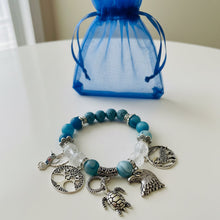 Load image into Gallery viewer, Bracelet with charms and blue beads on a white surface with a blue drawstring bag in the background.