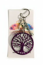 Load image into Gallery viewer, A purple glitter tree of life keychain with a silver metal tree design, attached to a silver split ring and keychain chain, displayed on a white branded backing card over a green patterned fabric background.