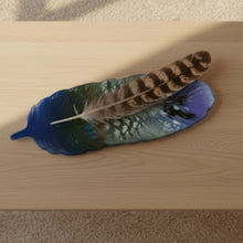 Load image into Gallery viewer, A decorative glass feather sculpture with a gradient design transitioning from purple and black at the top, through white and green, to a deep blue base, resting on a light brown surface.