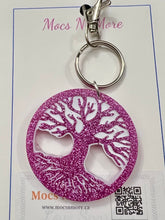 Load image into Gallery viewer, A pink glitter circular keychain with a white tree-of-life cutout design, attached to a silver metal split ring and clasp, displayed on a branded white card.