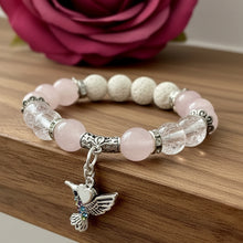Load image into Gallery viewer, A beaded stretch bracelet featuring pale pink and cream beads, clear crystal-like beads, and a dangling silver hummingbird charm with a small heart and iridescent accents, displayed on a dark surface.