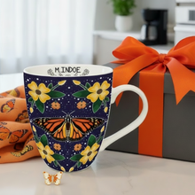 Load image into Gallery viewer, Mug with butterfly design and floral patterns next to a gift box with an orange ribbon.