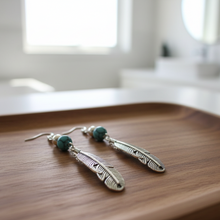 Load image into Gallery viewer, Feather-shaped earrings with green beads on a wooden tray.